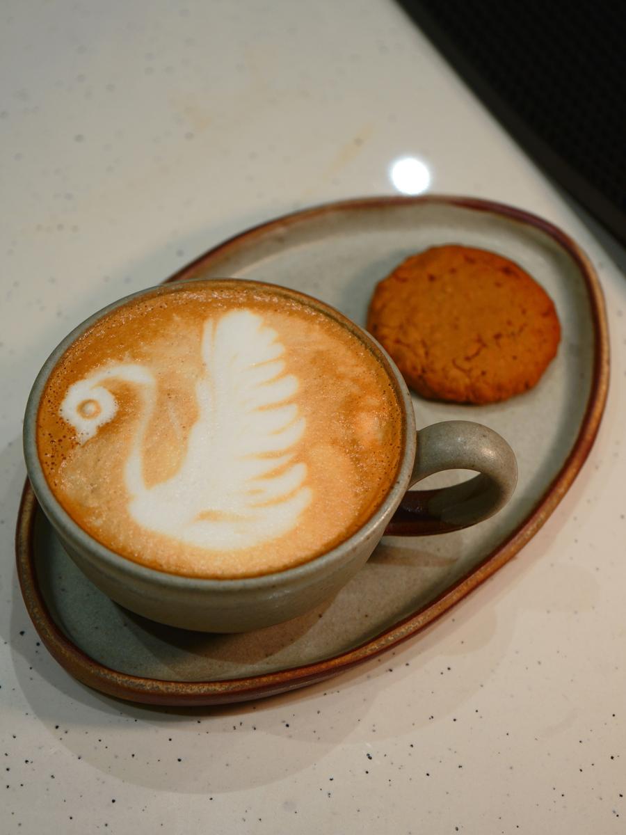 Swan latte art and a biscuit on a ceramic plate at Rithm Cafe